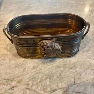 May include: A vintage brass oval-shaped container with a grape cluster and leaf design on the front. The container has two handles and a ribbed exterior. The brass has a warm, aged patina.