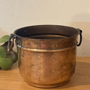 May include: A vintage copper bucket with a dark rim and two metal handles. The aged copper has a warm, reddish-brown hue, with visible patina and slight imperfections. The bucket sits on a wooden surface, suggesting a rustic or antique aesthetic.