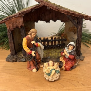 Può includere: Un presepe con le statuine di Giuseppe, Maria e Gesù bambino in una mangiatoia. Le figure sono dipinte con abiti e lineamenti dettagliati. La stalla ha un aspetto rustico e invecchiato, con un tetto in legno e muri in pietra.