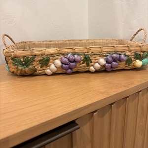May include: Rectangular woven basket with rope handles. The basket is decorated with clusters of purple and white grapes, green leaves, and small turquoise accents. The basket is made of natural-coloured woven material.