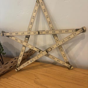 May include: A star-shaped wall decoration made from vintage wooden folding rulers. The rulers are beige with black markings and metal joints. The star is displayed on a light-colored wall and a wooden surface.