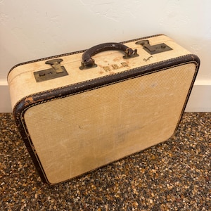 Könnte beinhalten: Vintage beiger Koffer mit braunen Lederbesatz und Griff. Der Koffer hat Metallverschlüsse und Eckschützer. Die Außenseite ist ein strukturiertes Gewebe. Der rechteckige Koffer steht auf einer gesprenkelten Oberfläche.