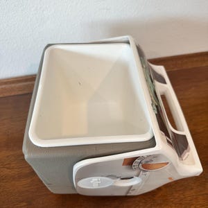 May include: A white and grey cooler with a handle and a white interior. The cooler has a grey base and a design on the side. The cooler is on a wooden surface.