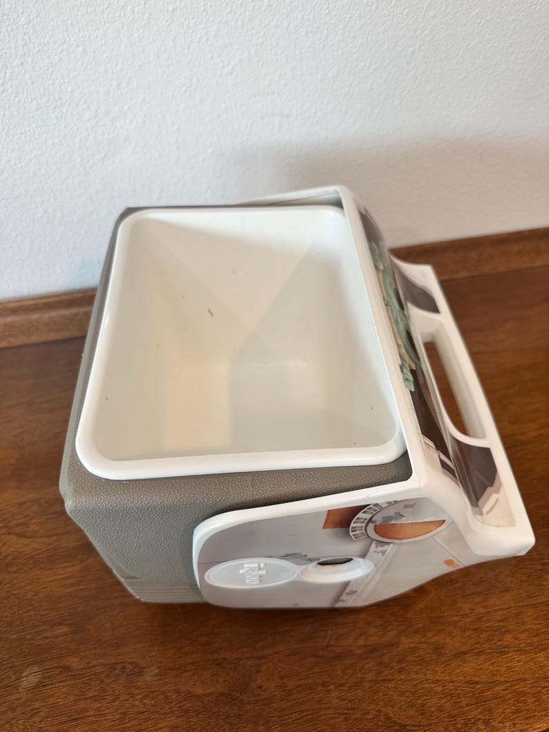 May include: A white and grey cooler with a handle and a white interior. The cooler has a grey base and a design on the side. The cooler is on a wooden surface.