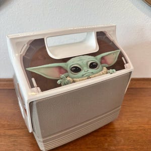 May include: A gray and white cooler featuring a Baby Yoda character peeking out from the top. The cooler has a handle and a clear plastic top. The Baby Yoda figure is green with large eyes and pointed ears.