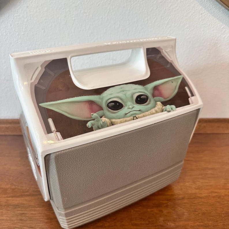 May include: A grey and white cooler featuring a Baby Yoda character peeking out from the top. The cooler has a handle and a clear plastic top. The Baby Yoda figure is green with large eyes and pointed ears.
