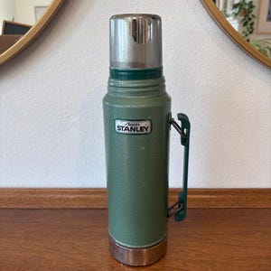 May include: A vintage green Stanley flask with a silver lid and base. The flask has a green handle and the "Stanley" logo is visible on the front. The flask is on a wooden surface.