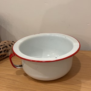 Puede incluir: Orinal de esmalte blanco con borde y asa rojos. El orinal tiene forma redondeada y una abertura ancha. El asa es de color rojo brillante.