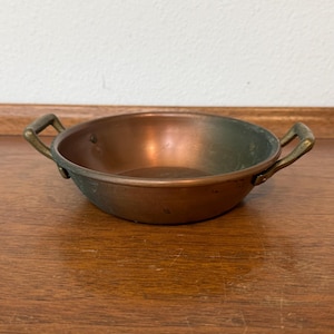 May include: A shallow, round copper pan with a polished interior and a weathered, darkened exterior. The pan features two small, curved brass handles attached to opposite sides with visible rivets, creating a functional and simple design.