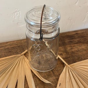 May include: A clear glass Ball wide mouth jar with a wire bail closure. The jar has a textured surface and the word "Ball" is embossed on the front.