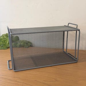 May include: A rectangular gray metal mesh storage shelf featuring a two-tier design with a flat top surface and a bottom base. The unit is constructed from a fine wire grid material and includes vertical support bars on each side for stability.