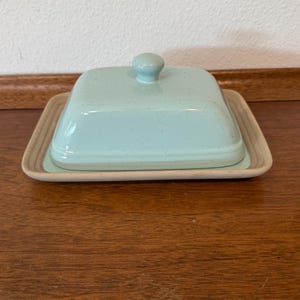 May include: A rectangular butter dish featuring a light mint green lid with a speckled finish and a knob handle, resting on a solid beige base with a raised rim. The ceramic container has a smooth, glossy surface and a clean, functional design.
