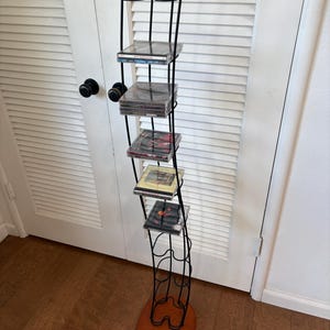 May include: A tall, slender CD storage rack featuring a black metal wire frame with a curved, wave-like design. The unit has a solid wooden base and four small wooden spheres at the top. It holds several CD cases stacked on horizontal wire shelves.