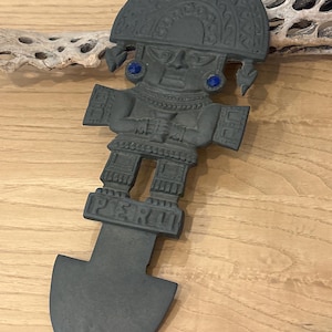 May include: A dark gray metal decorative figure in the shape of an Incan warrior. The figure has a large headdress, blue stone accents, and the word "PERU" at the base. The figure has a shovel-like shape at the bottom.