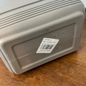 May include: A grey Igloo cooler with a textured surface and horizontal ridges. The cooler has a white top and a label with text. The cooler is on a wooden surface.