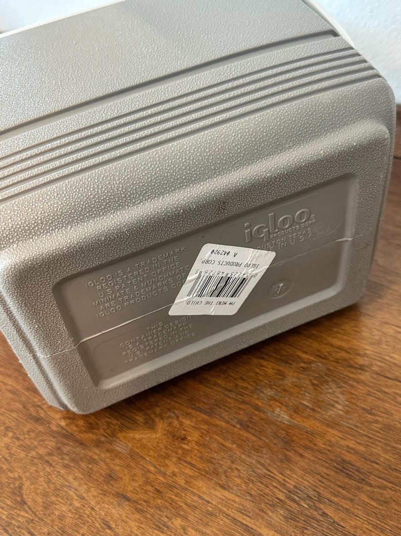 May include: A grey Igloo cooler with a textured surface and horizontal ridges. The cooler has a white top and a label with text. The cooler is on a wooden surface.