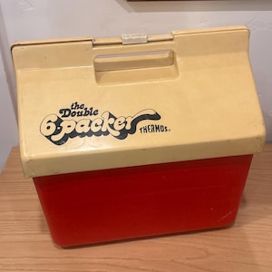 Puede incluir: Nevera Thermos "Double 6-Packer" vintage roja y crema. La nevera tiene una tapa color crema con un asa negra y las palabras "the Double 6-Packer THERMOS" impresas en la parte delantera. La base es roja.
