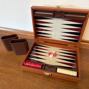 May include: A wooden backgammon set with a brown exterior, open to reveal a playing surface with black, white, and burgundy triangles. Red and white game pieces and dice are visible. Two brown dice cups are on the left.