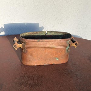 May include: An oval copper container with a horizontal ribbed design, featuring two brass side handles with dark wooden grips. The metal surface shows signs of oxidation and patina, with visible green verdigris accents along the rim and handles.