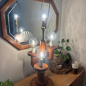 May include: A multi-tiered wooden table lamp featuring a central vertical post with three extending arms, each holding a clear round light bulb. The lamp has a circular wooden base and a brass-colored metal harp at the top for a shade.