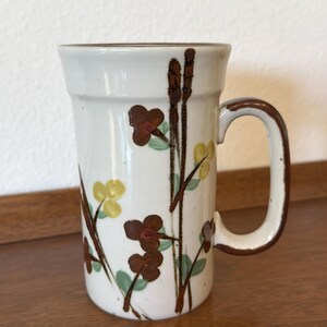 May include: A white ceramic mug with a brown handle and rim. The mug is decorated with brown, yellow, and green floral designs. The mug is sitting on a wooden surface.