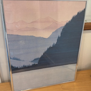 May include: A rectangular wall art print in a thin silver metal frame featuring a landscape illustration of layered mountains in shades of pale pink and blue, with a dark blue forest silhouette on the right side and a light gray foreground.