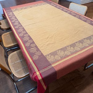 May include: A rectangular tablecloth featuring a large solid tan center panel bordered by a wide band of dark maroon fabric decorated with a repeating pattern of light-colored maple leaves, finished with a thin yellow stripe and red edge.