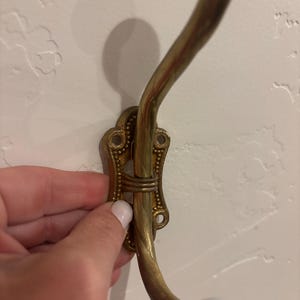 May include: A brass wall hook featuring an ornate, rectangular mounting plate with decorative beaded edges and four screw holes. The hook has a curved, twisted metal arm that extends outward and terminates in a smooth, round white ceramic knob.