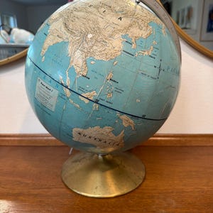May include: A Rand Mark I globe featuring a light blue ocean and tan landmasses. The globe is mounted on a circular brass-colored metal base and includes a silver-colored metal meridian ring with degree markings. Text on the globe includes Rand Mark I.