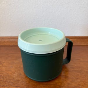 May include: A dark green travel mug with a light green lid and a black handle. The mug is cylindrical and appears to be made of plastic. The lid has a small hole in the center. The mug is sitting on a wooden surface.