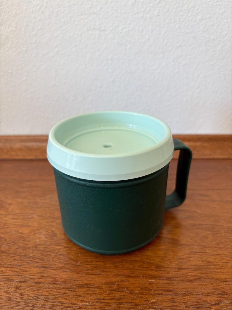 May include: A dark green travel mug with a light green lid and a black handle. The mug is cylindrical and appears to be made of plastic. The lid has a small hole in the center. The mug is sitting on a wooden surface.