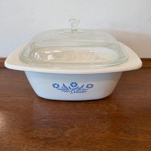 May include: A white square casserole dish with rounded corners and a clear glass lid. The exterior features a blue floral pattern centered on the front. The dish has wide, flat handles on opposite sides and a small glass knob on the lid.