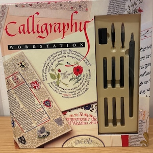 May include: A calligraphy workstation set in a box. The box features the word "Calligraphy" in red script, with the text "WORKSTATION" below. Inside, a selection of black calligraphy pens and nibs are visible. The box has decorative floral and text elements.