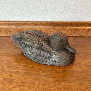 May include: A detailed sculpture of a duck with textured feathers. The duck has a brown head with a green stripe and a dark gray beak. The body is primarily gray with detailed feathering.