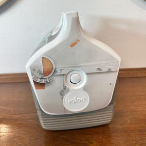 May include: White and grey Igloo cooler with a push-button lid and a handle. The cooler has a design with orange and grey accents. The Igloo logo is visible on the front of the cooler.