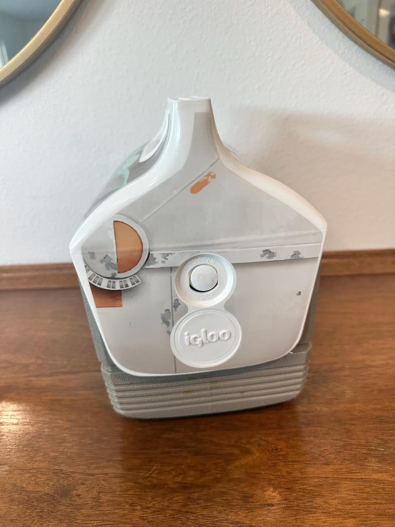 May include: White and grey Igloo cooler with a push-button lid and a handle. The cooler has a design with orange and grey accents. The Igloo logo is visible on the front of the cooler.