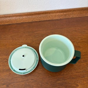 May include: A green travel mug with a matching lid. The mug has a dark green exterior and a light green interior. The lid has a small opening for drinking. The mug is sitting on a wooden surface.