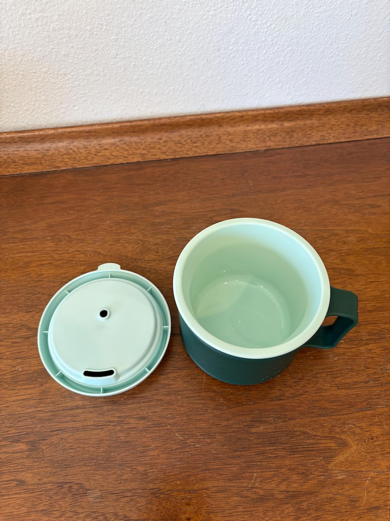 May include: A green travel mug with a matching lid. The mug has a dark green exterior and a light green interior. The lid has a small opening for drinking. The mug is sitting on a wooden surface.