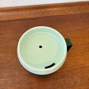 May include: A green and white travel mug with a dark green handle. The lid is light green with a small hole and a curved opening. The mug is on a wooden surface.