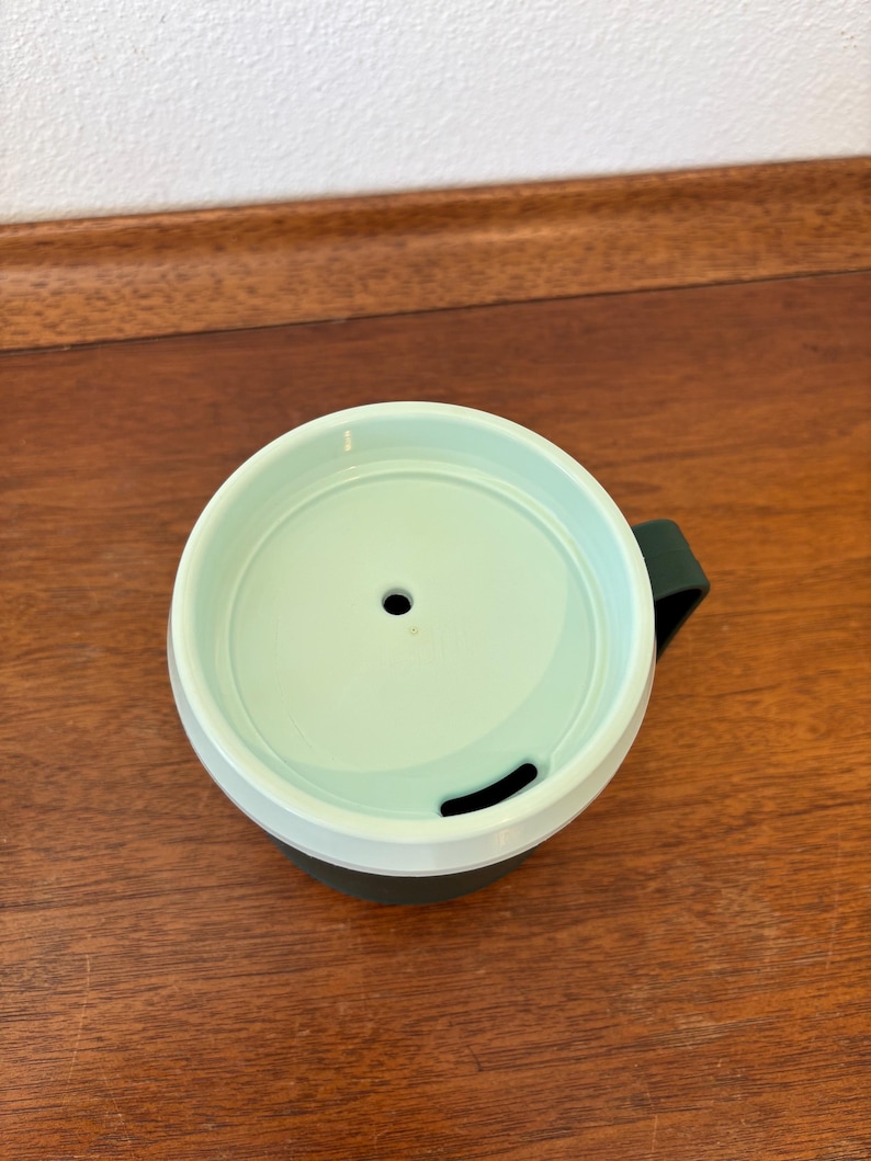 May include: A green and white travel mug with a dark green handle. The lid is light green with a small hole and a curved opening. The mug is on a wooden surface.