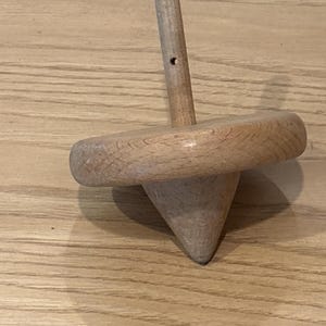 May include: A wooden spinning top with a conical base, a circular midsection, and a cylindrical handle. The toy is made of light-colored wood and sits on a wooden surface. The top has a small hole in the handle.