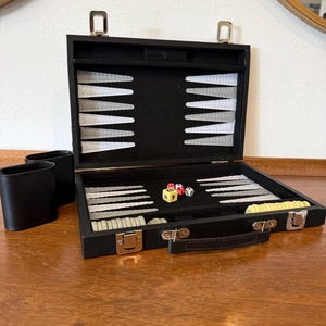 May include: A black backgammon set in a briefcase-style case with a black felt interior, white and grey triangular points, white and yellow circular checkers, two black dice cups, and three dice, including one large yellow doubling cube with numbers.