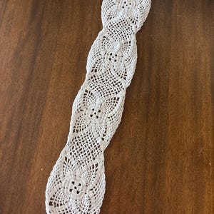 May include: A white, handmade crochet table runner with a floral design. The runner is long and narrow, with a repeating pattern of openwork flowers and a scalloped edge. The runner is displayed on a dark brown wooden surface.