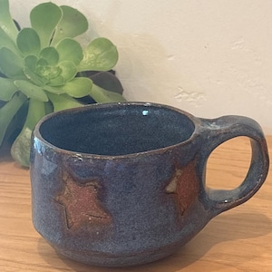 May include: A handmade ceramic mug with a dark blue glaze and two embossed star designs in a reddish-brown color. The mug has a rounded shape and a sturdy handle, perfect for enjoying hot beverages. The mug is approximately 3.5 inches in diameter.