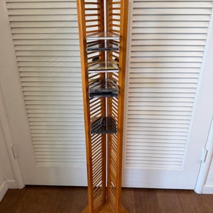 May include: A tall, light brown wooden media storage tower with a triangular base and top. The unit features vertical slats on three sides and horizontal shelves designed to hold compact discs. Several CD cases are stacked on the internal shelves.