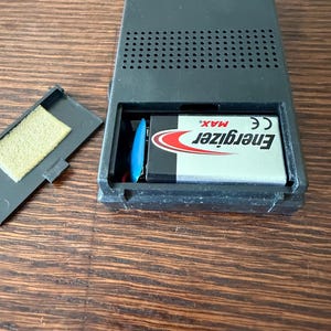 May include: Black Emerson radio with an open battery compartment revealing an Energizer Max battery. The radio has a speaker grill and text indicating AM/FM frequencies. The model number is P3761.