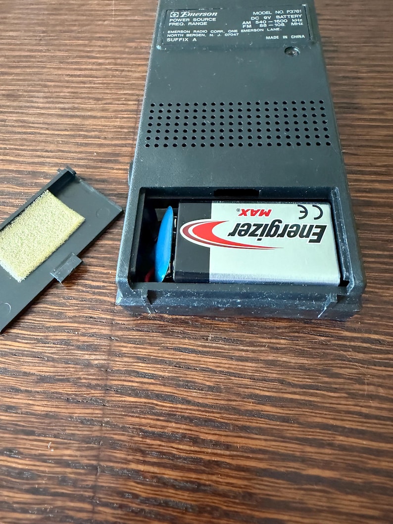 May include: Black Emerson radio with an open battery compartment revealing an Energizer Max battery. The radio has a speaker grill and text indicating AM/FM frequencies. The model number is P3761.