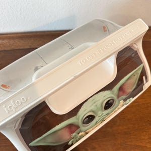 May include: White Igloo lunchbox featuring a graphic of Grogu, also known as Baby Yoda, from The Mandalorian. The lunchbox has a white handle and the words "TO OPEN PUSH BUTTON" printed on the lid.