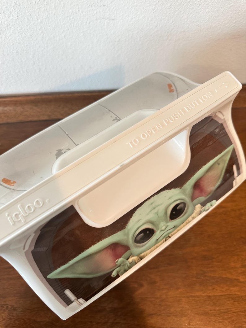 May include: White Igloo lunchbox featuring a graphic of Grogu, also known as Baby Yoda, from The Mandalorian. The lunchbox has a white handle and the words "TO OPEN PUSH BUTTON" printed on the lid.