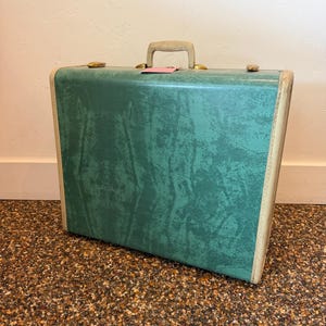 May include: Vintage green suitcase with a textured surface, brass hardware, and a light-colored handle. The suitcase has a rectangular shape with rounded corners and a cream-colored trim. A pink tag hangs from the handle.